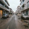 Calle de ciudad cubierta de lodo tras una riada, con dos coches aparcados muy sucios y edificios de apartamentos a ambos lados bajo un cielo gris.