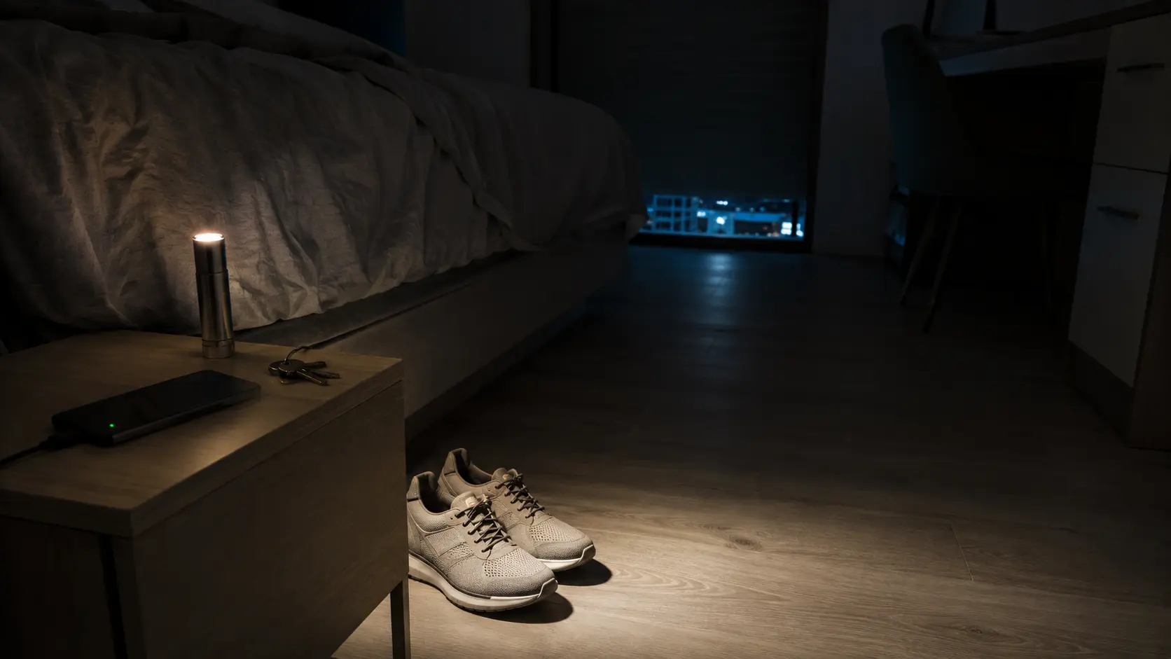 Mesilla de noche con una linterna encendida y unas zapatillas cerradas debajo de la cama